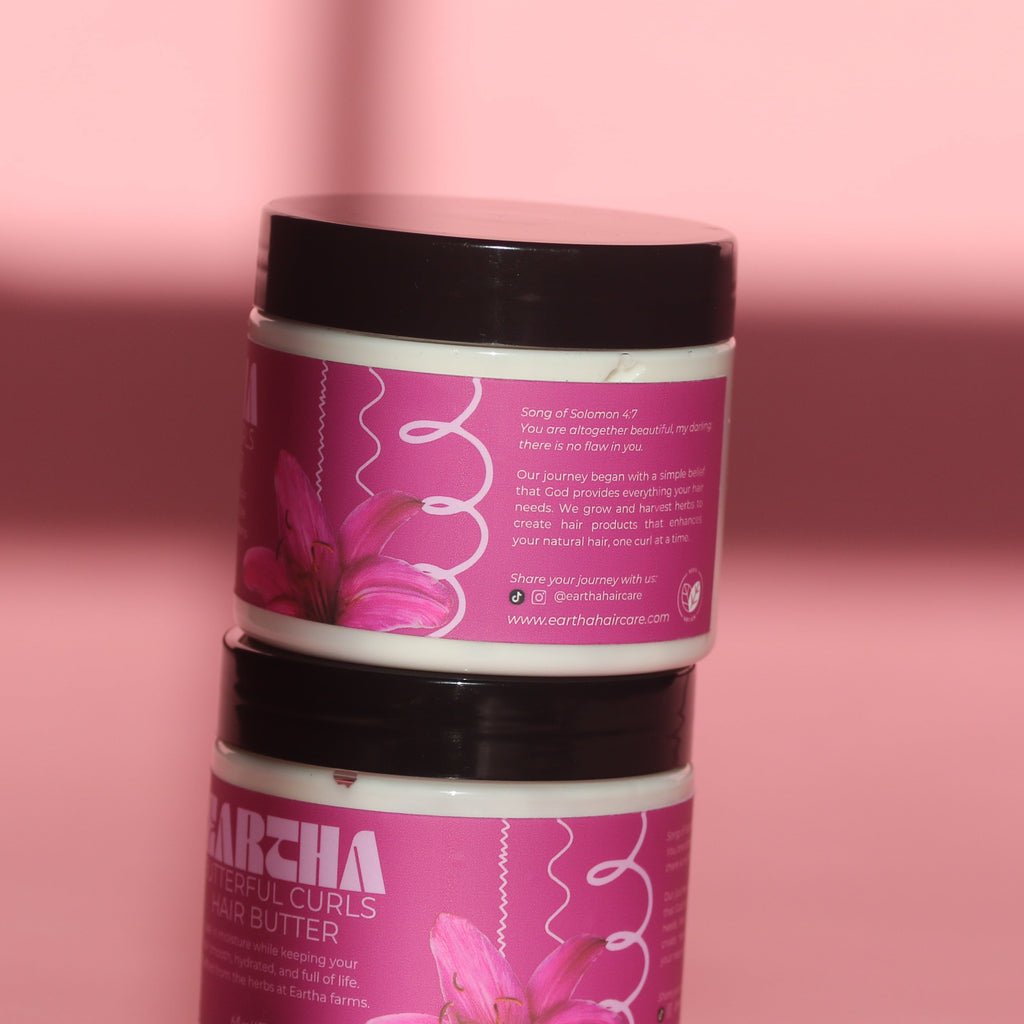 Two stacked white plastic jars of hair oil with black lids and pink labels featuring a pink flower design, set against a solid pink background.